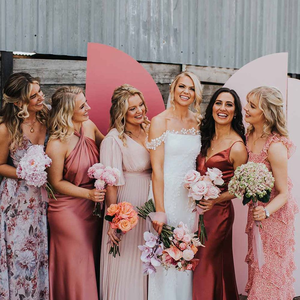 Bride wearing lace off the shoulder wedding dress standing in front of pink wedding screen backdrop with bridesmaids in mismatched bridesmaid dresses