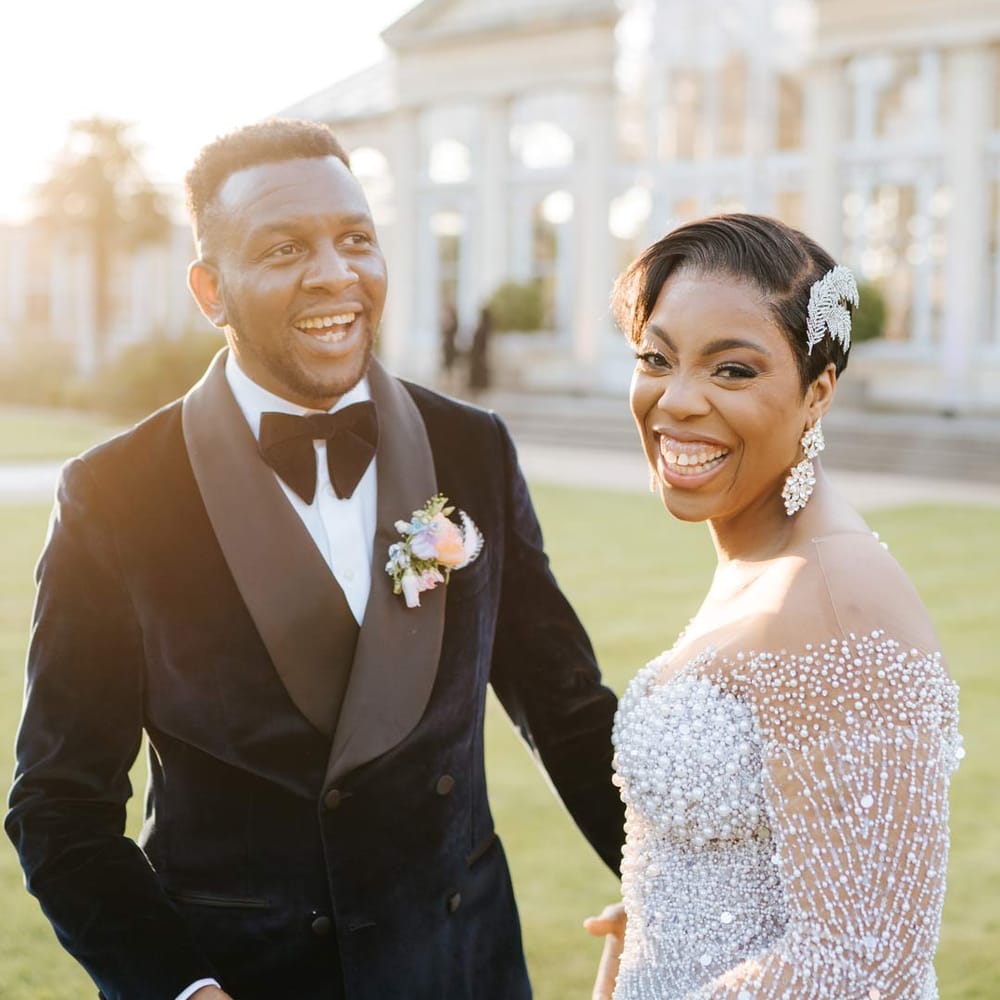 Groom in velvet midnight blue double breasted groom’s blazer, velvet bowtie and peach garden rose boutonniere with bride in illusion v neck sheer off shoulder long sleeve pearl embellished wedding gown at Syon Park vow renewal
