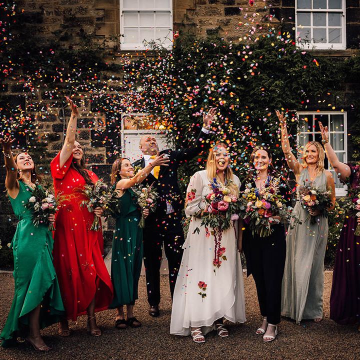 Bridesmaids wearing mismatched bright and colourful dresses throw confetti over the brides 