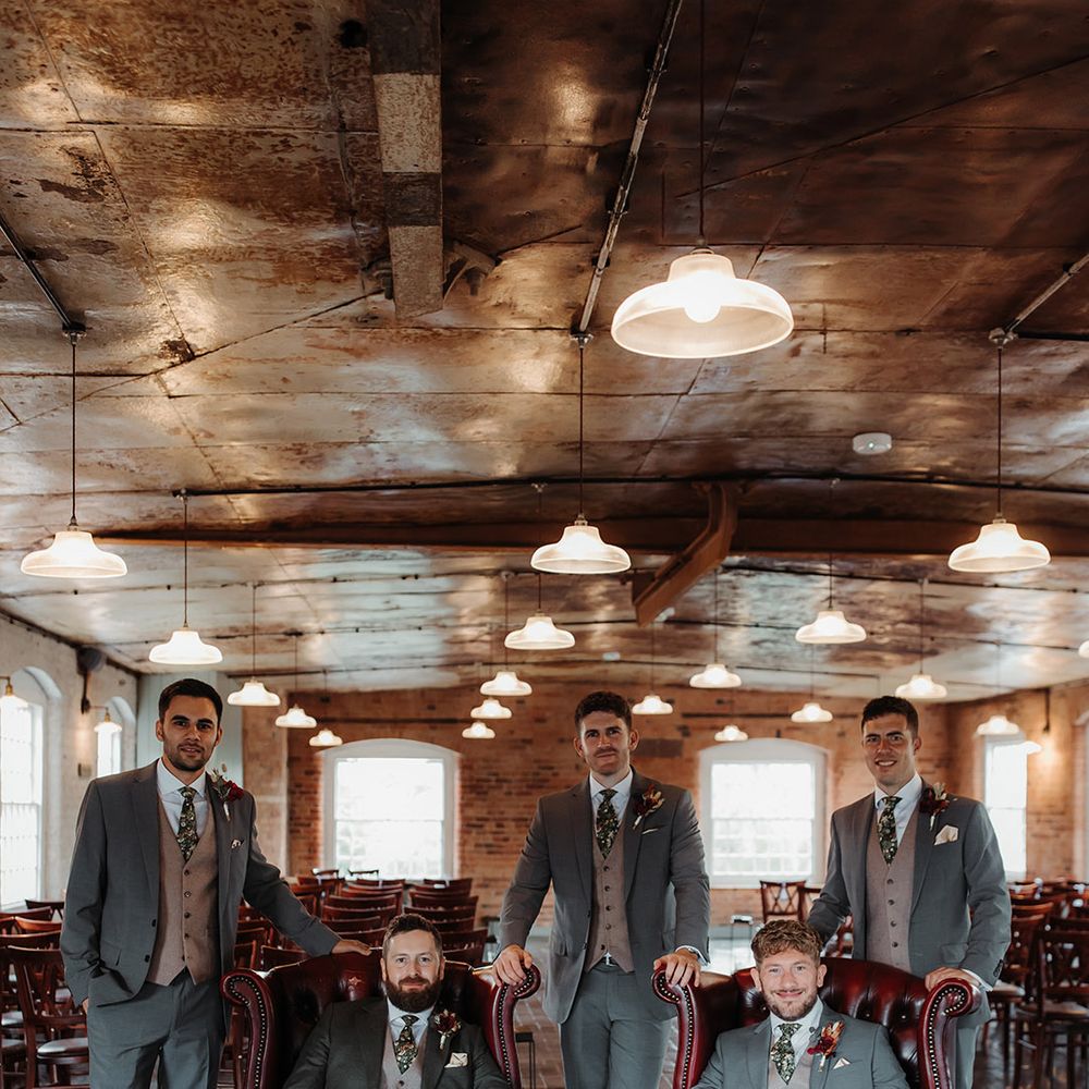 The groom and groomsmen pose together for a group photo with the groomsmen posing on red leather chairs 