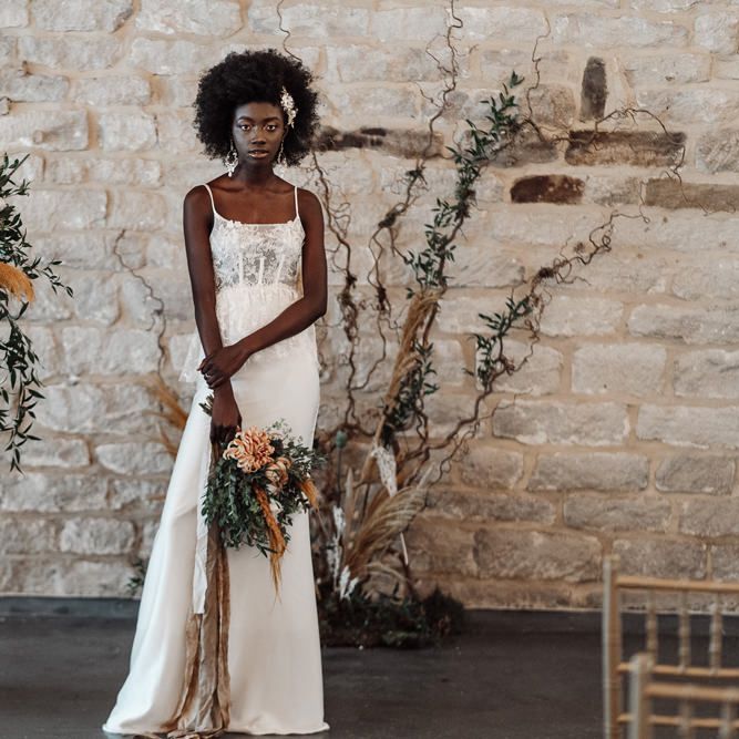 Black bride with afro hair in a lace peplum wedding dress standing by a pillar with orange and green flowers at Ponden Mill