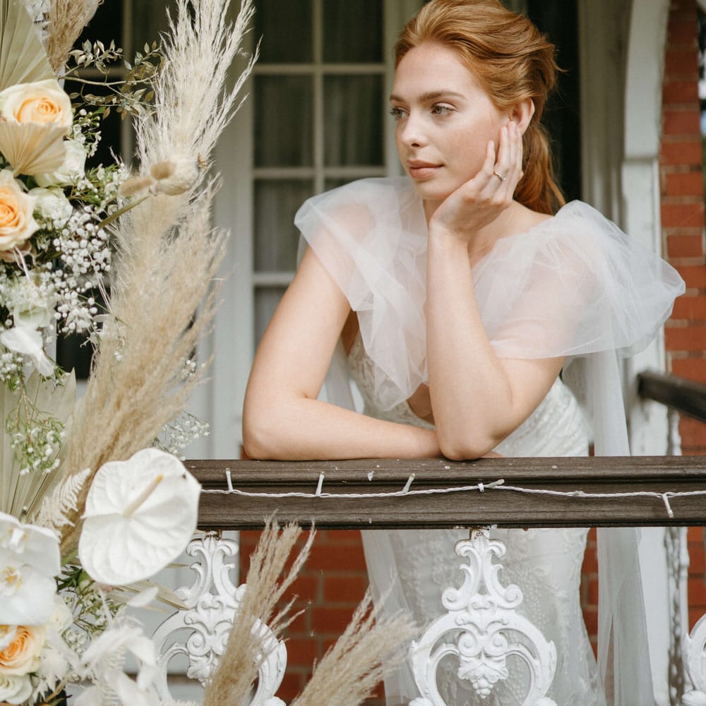 Bride leaning on wooden railing at Berwick Lodge, wearing a puff tulle shoulder detail wedding dress, next to a boho dried flower arch