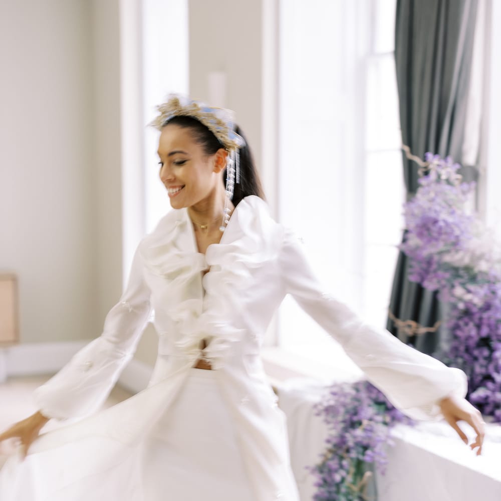 Bride holds out wedding gown with ruffled front whilst stood by lavender trailing from the window