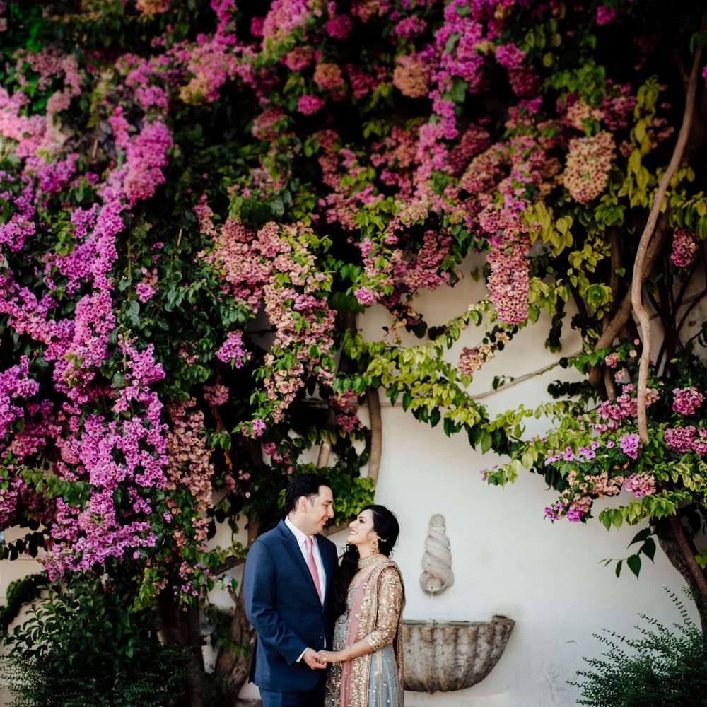 Bride & groom look lovingly at one another as they stand in front of bright pink floral tree as bride wears colourful saree