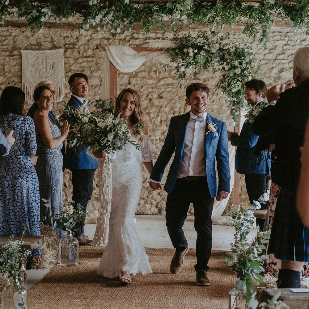 Bride in Grace Loves Lace bardot wedding dress with white and green wedding bouquet walks back down the aisle holding hands with groom in blue suit jacket and floral buttonhole for Isle of Wight wedding with macrame wedding decor