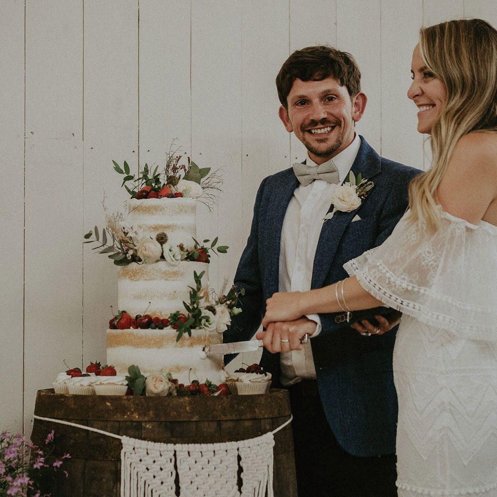 Bride in Grace Loves Lace bardot wedding dress and veils cuts three tier semi naked wedding cad with groom in blue suit jacket with floral buttonhole at Isle fo Wight wedding with macrame wedding decor