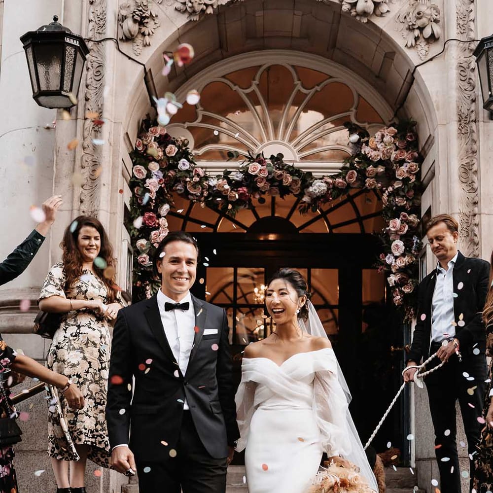 Groom in black suit with white shirt and black bow tie holding hands with Bride in silk and chiffon puff sleeve dress as wedding guests cheer and throw colourful confetti 