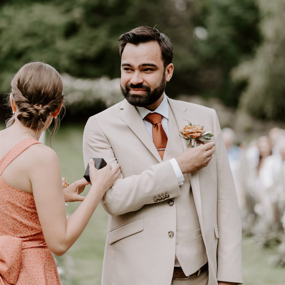 Groom in three piece beige Moss Bros. suit with floral buttonhole waits for bride with bridesmaid in orange dress during DIY garden wedding ceremony in Bedfordshire