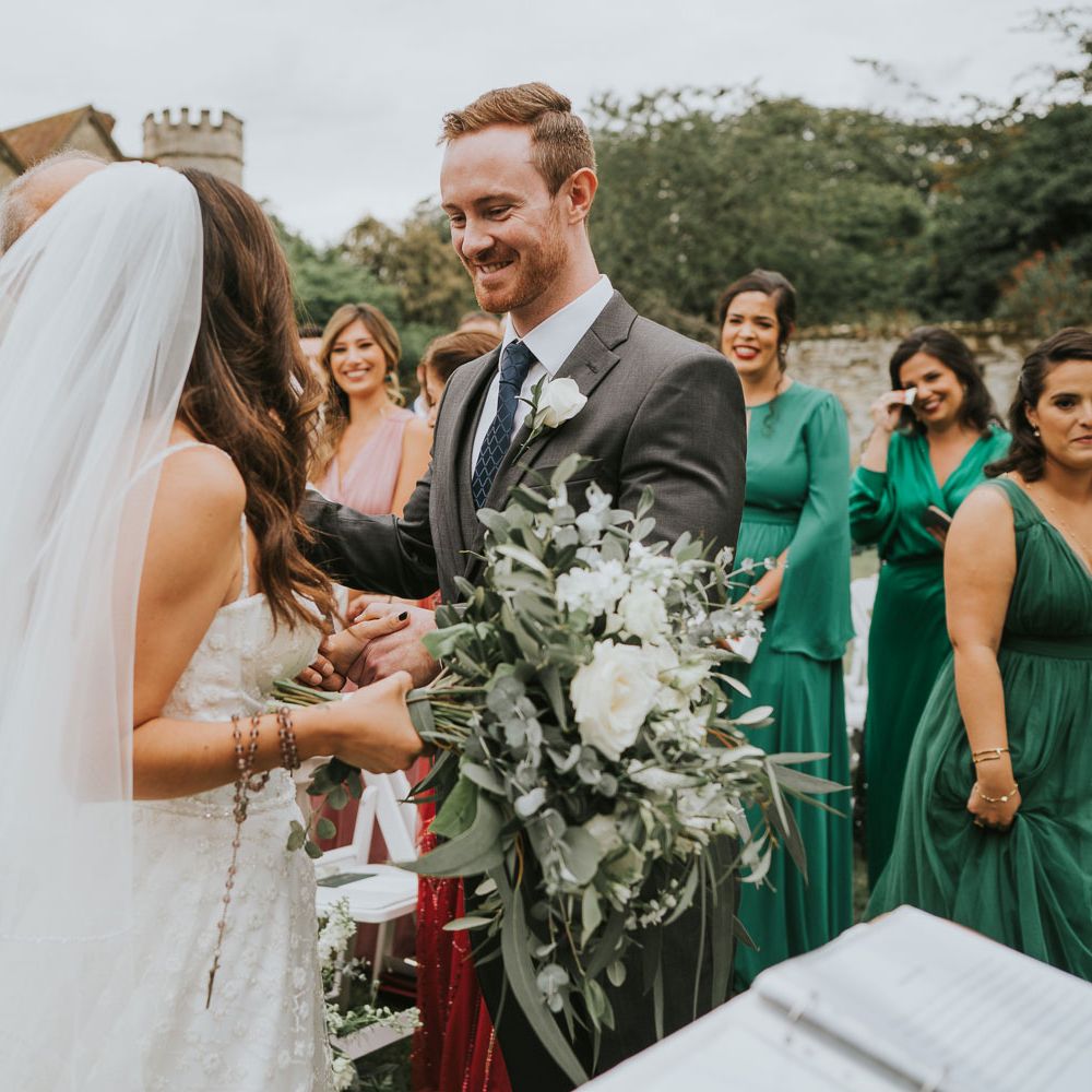 Bride in Pronovias wedding dress and veil holding white rose and green bridal bouquet stands with smiling groom in three piece grey suit during wedding ceremony 