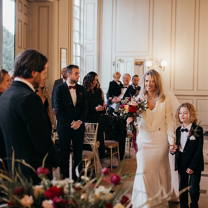 Bride walks down the aisle with son who wears black tie as bride wears Zara jacket and Suzanne Neville wedding gown 