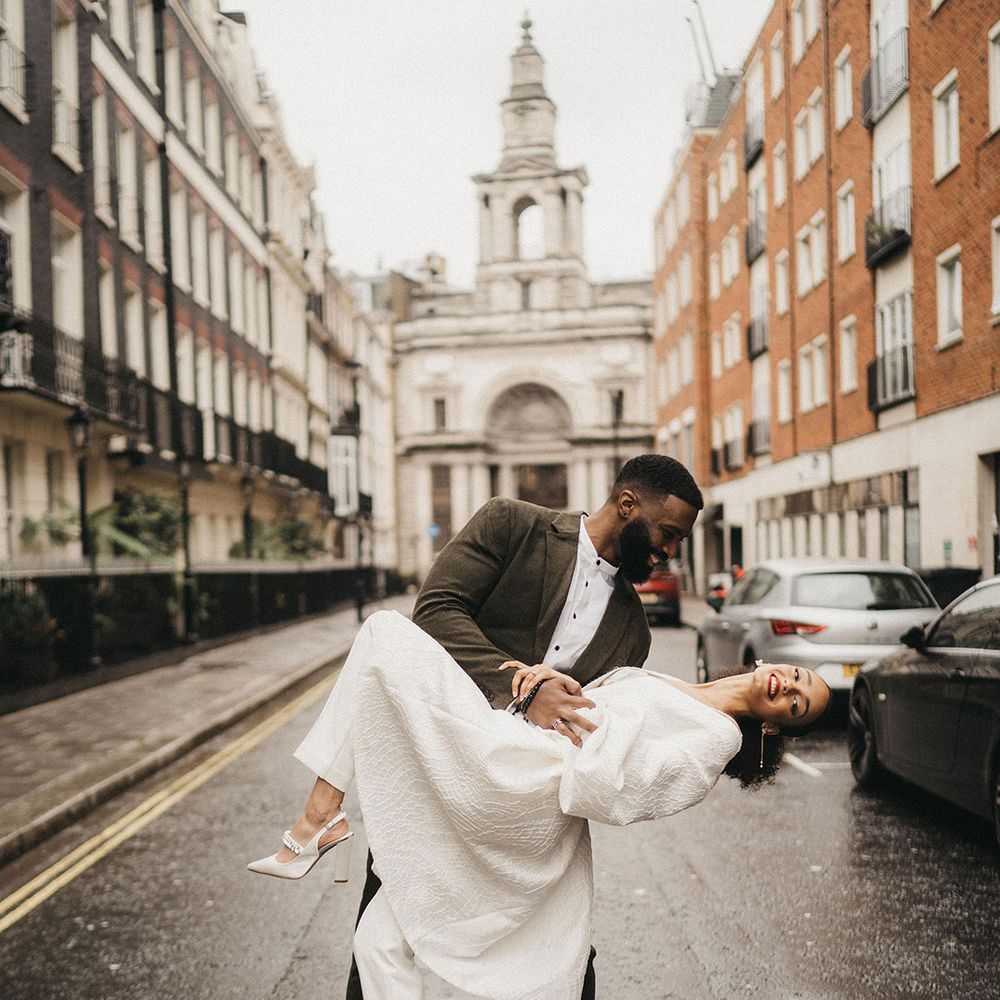 Black groom in a sage green wool wedding suit leaning his bride over for a kiss in a trouser suit and bridal coat in London 