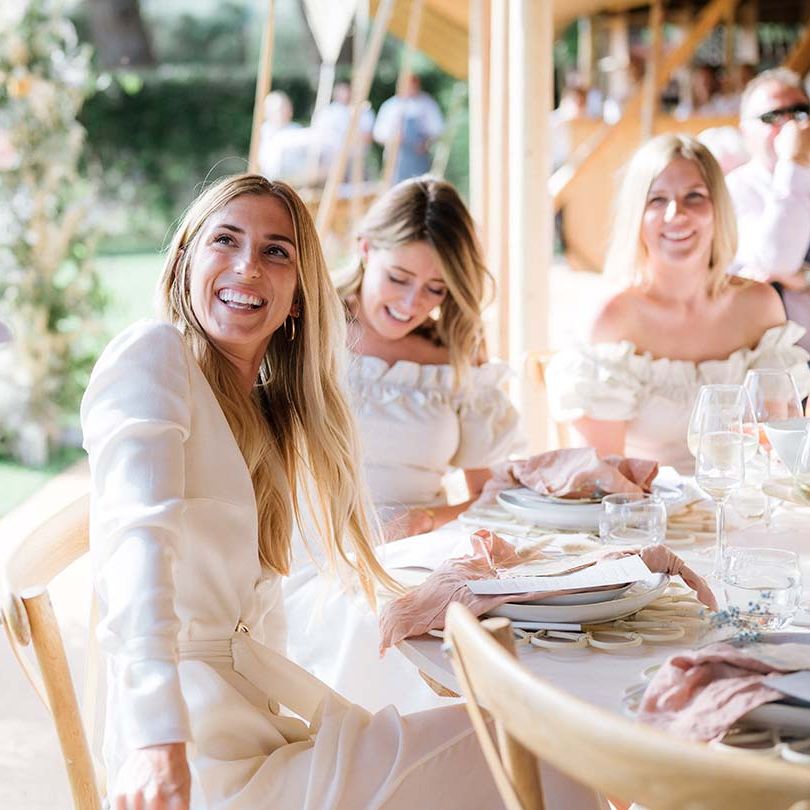 Bride in bespoke Bon Bride wedding dress with long sleeves and a belt smiles at the groom as he gives his speech