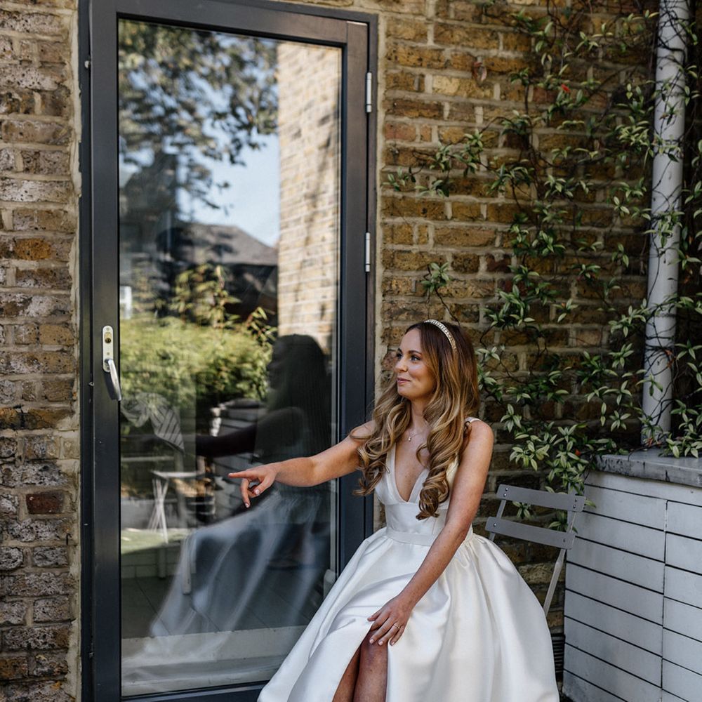 Bride with long wavy hair wearing a pearl headband sitting on a chair putting on her grey Manolo Blahnik wedding shoes 