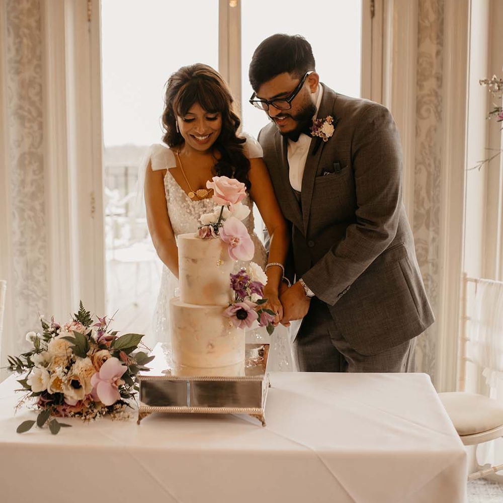 Bride and groom cutting into two tier neutral coloured wedding cake with mixed pinks and floral decorations