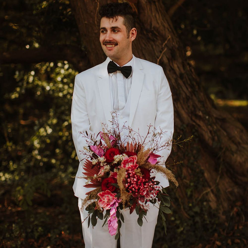 Groom in a three piece white groom suit with black bow tie holding a Brian red and pink wedding bouquet 