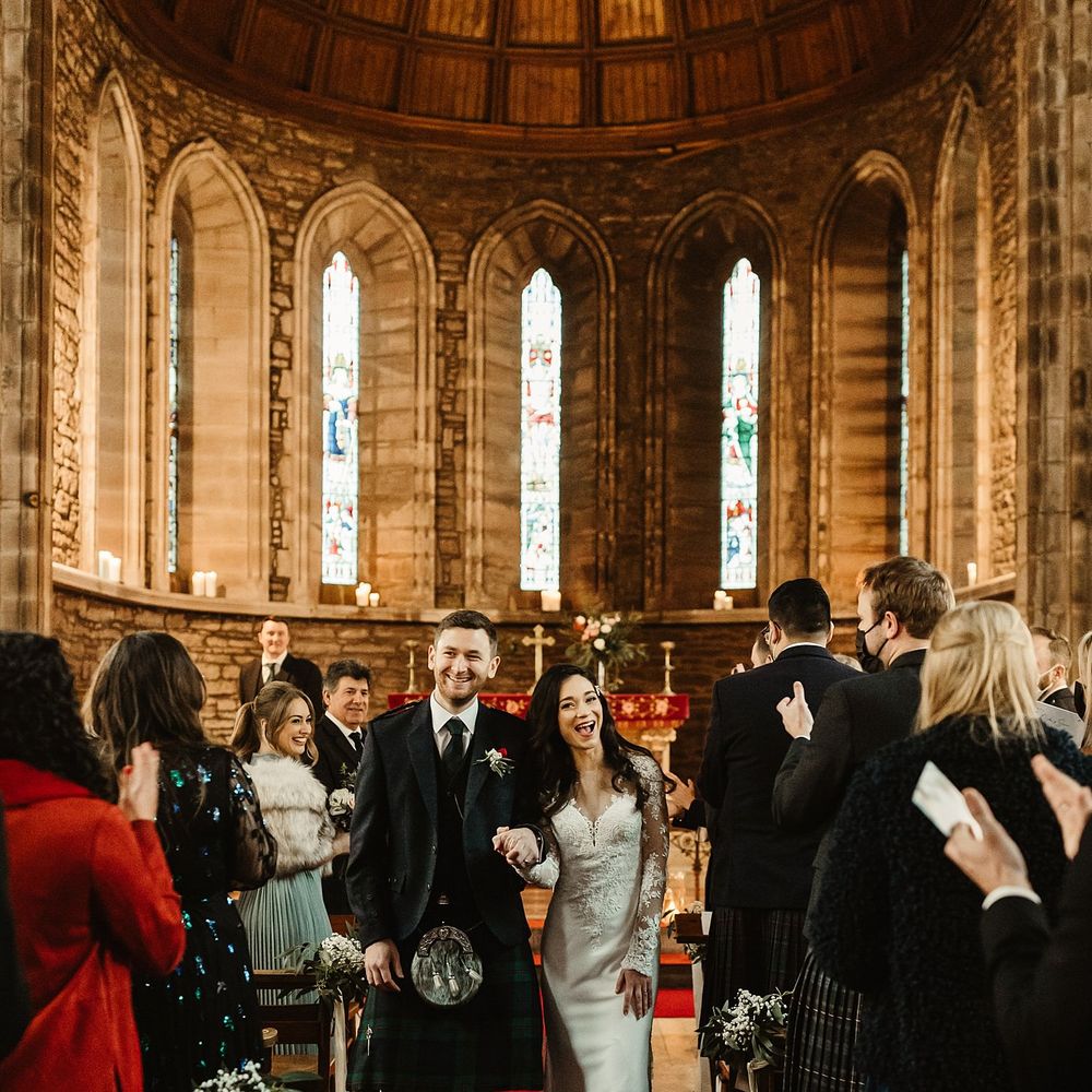 Bride wears lace wedding gown complete with long sleeves as she walks down the aisle with groom who wears kilt 
