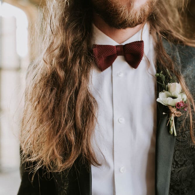 Groom with long hair in a jacquard black tuxedo and burgundy bow tie 