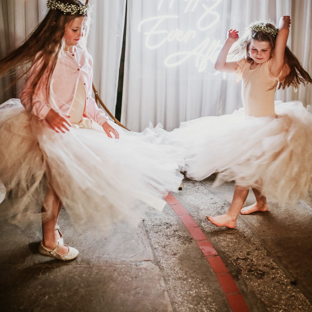 Flower girls wearing ballerina dresses and floral crowns dance in front of neon wedding sign
