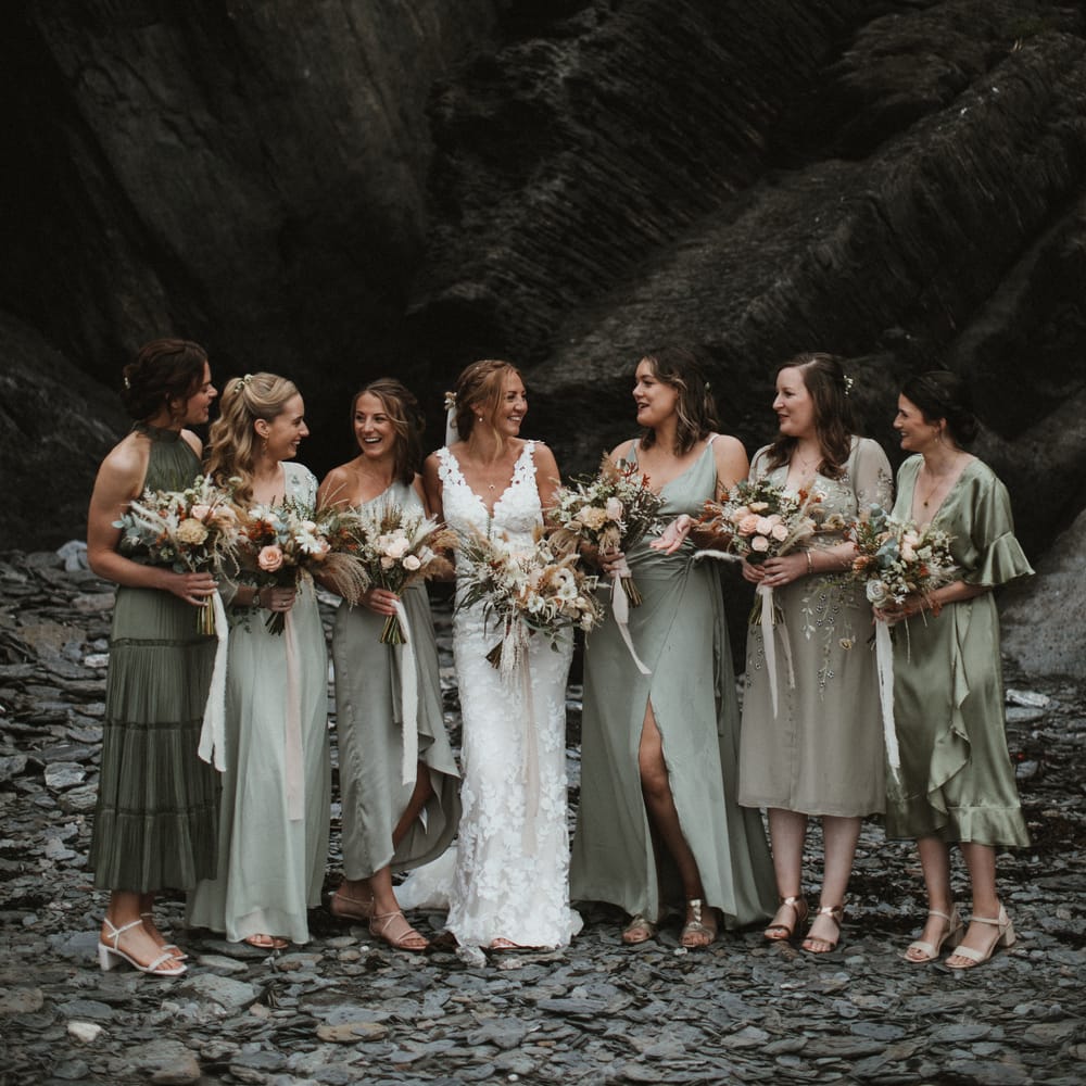 Bride stands with her bridesmaids who wear different shades of green bridesmaid dresses in differing styles as they hold champagne and rust coloured bouquets