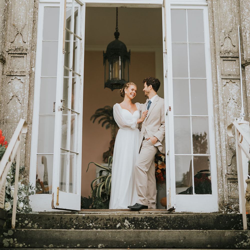 Groom in beige suit leans against the door frame as the bride puts a hand on his chest for Sezincote House wedding 