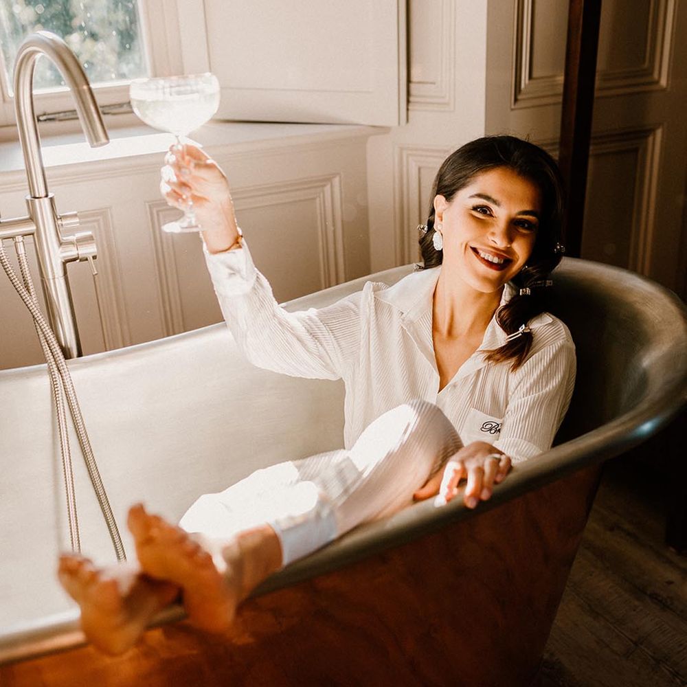 Bride lays out in a copper bath drinking champagne in white bridal pyjamas 