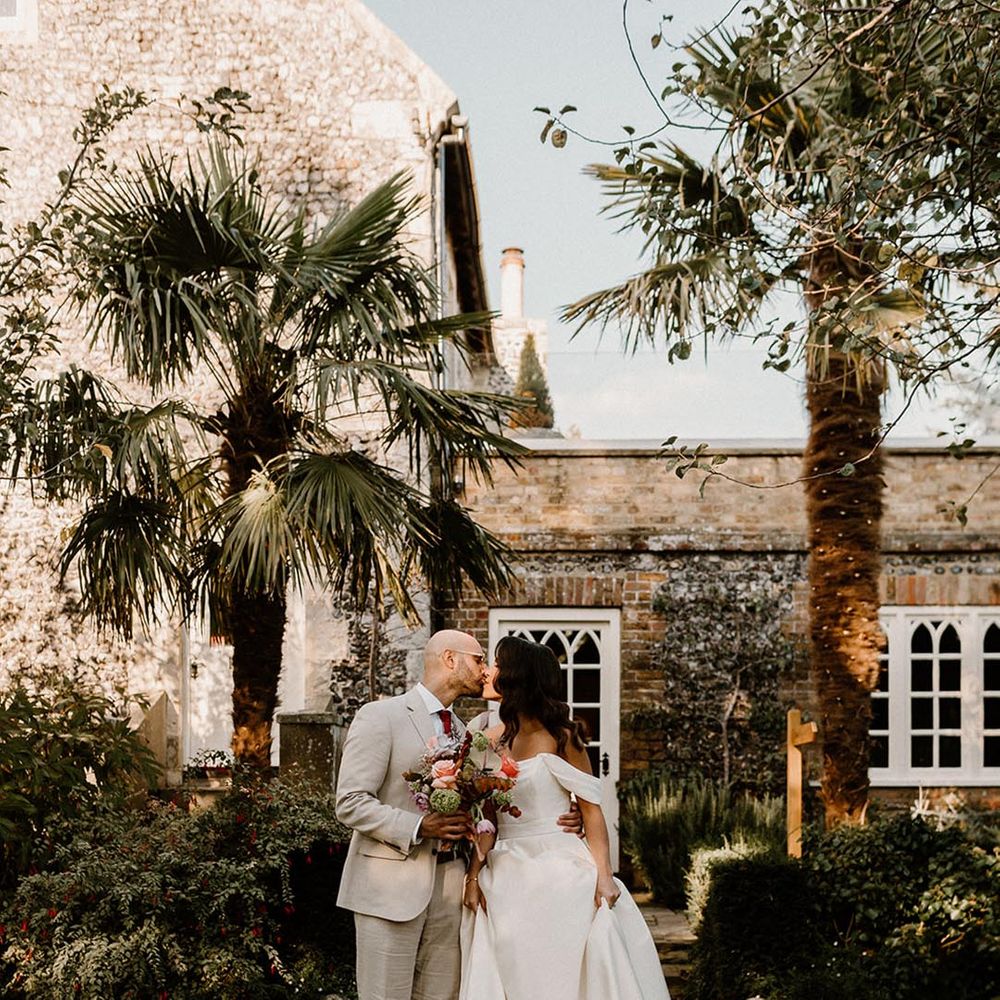 Groom in a pale suit with a dark red tie kisses the bride in an off the shoulder wedding dress for their autumn wedding 