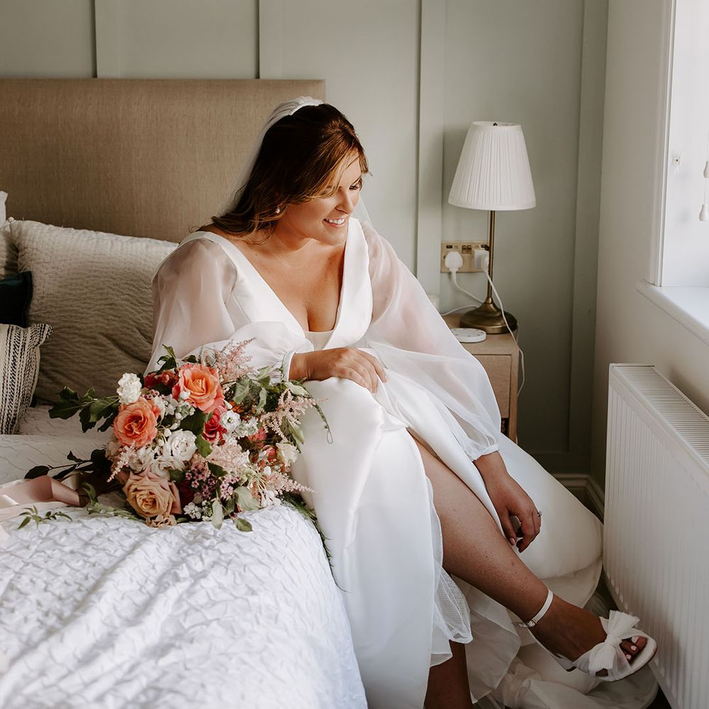 Bride wears full sheer sleeved romantic wedding dress whilst putting on strappy white bridal shoes with bow 