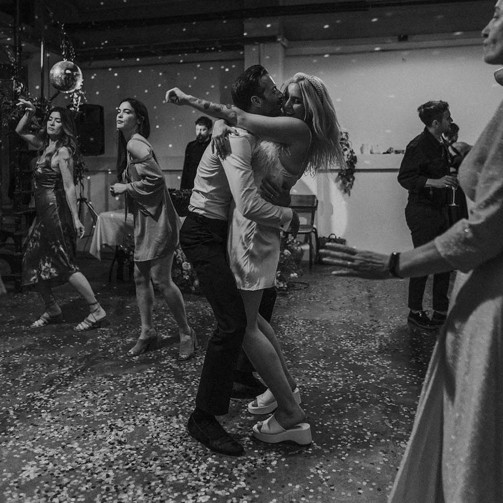 Bride in short satin dress with feather trimmed neckline and groom in dark tux kissing and dancing at Hackney Studios