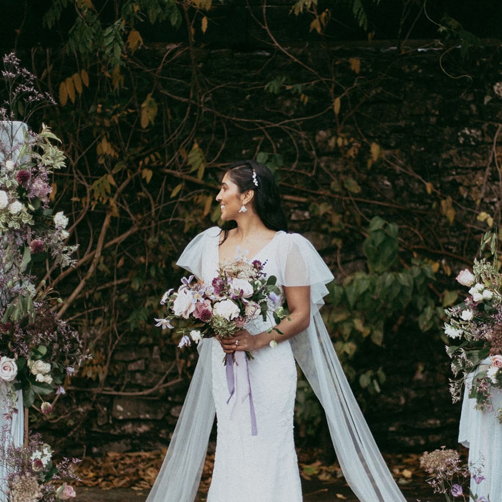 Bride in lace wedding dress with puff lace sleeves and bridal wings holding mixed floral bouquet with with white carnations, purple tulips, roses and foliage