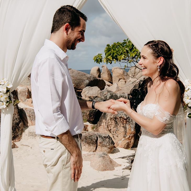 Groom in crisp white shirt and light coloured chinos belt putting wedding ring on bride wearing off the shoulder lace wedding dress 