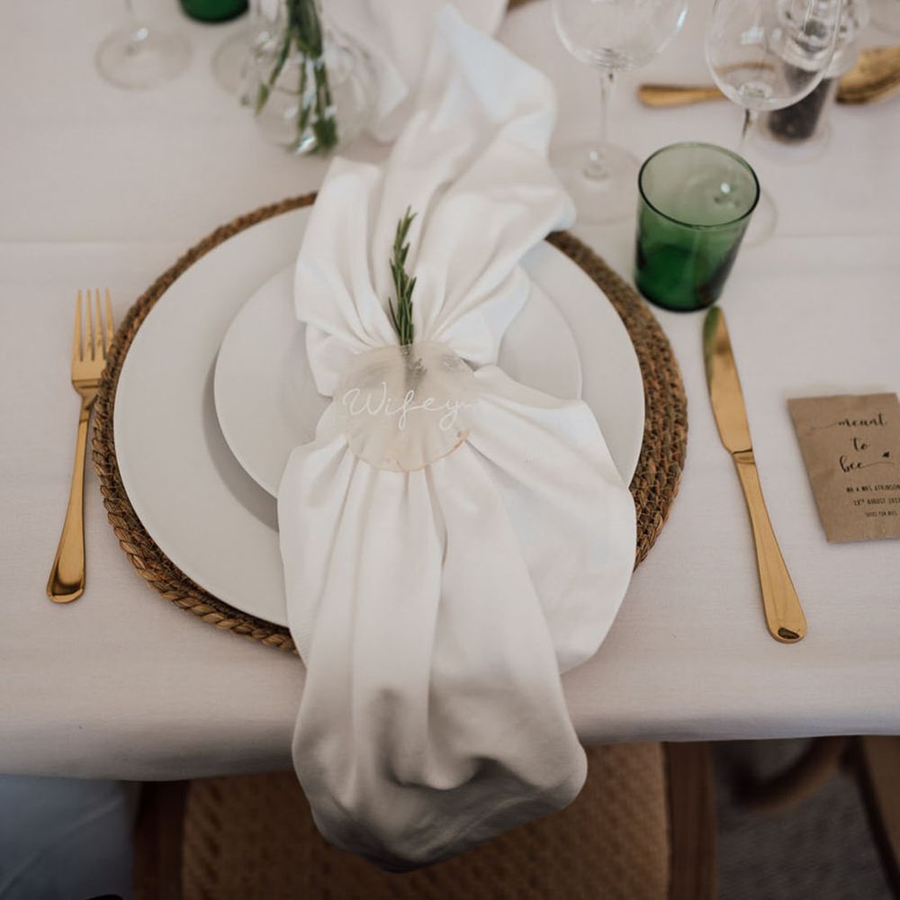 Place setting with wicker place mat, white plates, green coloured glassware, gold cutlery, with white napkin and frosted place name 