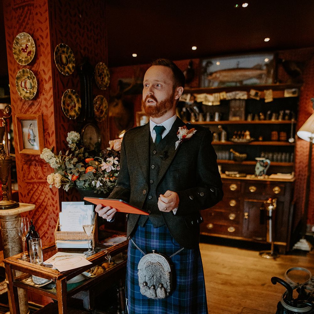 Best man wearing dark grey blazer and waistcoat, blue tie, autumnal boutonniere and dark blue kilt doing wedding speech