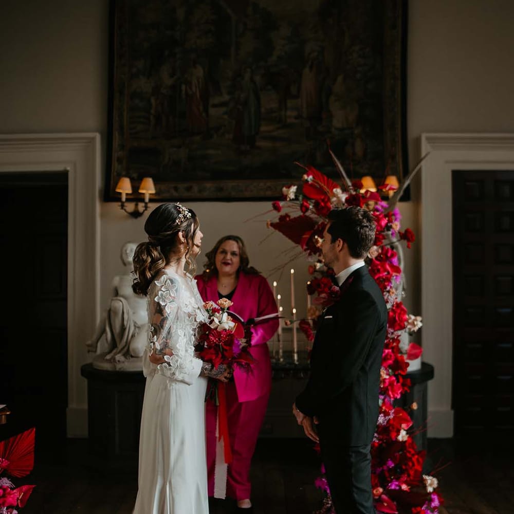 Wedding celebrant in hot pink suit speaking to bride and groom at the alter of Elmore Court with large standing floral decor with dahlias, orchids, roses, amaryllis, anthuriums and dried flowers 