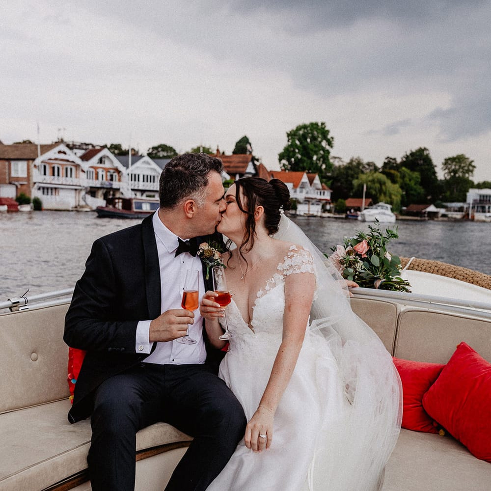Groom in black tie kissing the bride in a flower lace wedding dress as the drink alcohol and take a boat ride around the Thames 