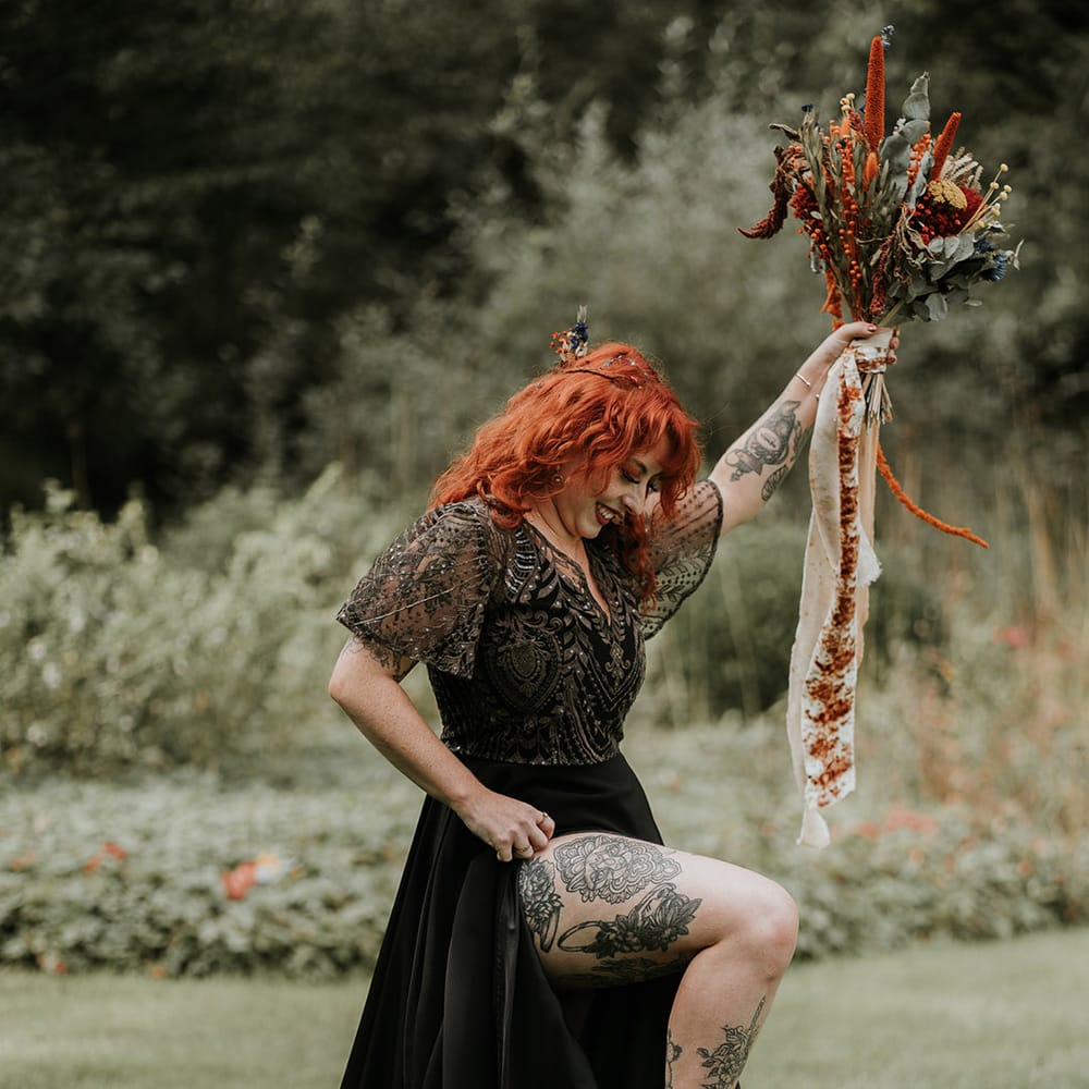 Bride in embellished v neck short sleeve black wedding dress showing off her tattoos holding large burnt orange and eucalyptus dried flower bouquet with orange and off white ribbons