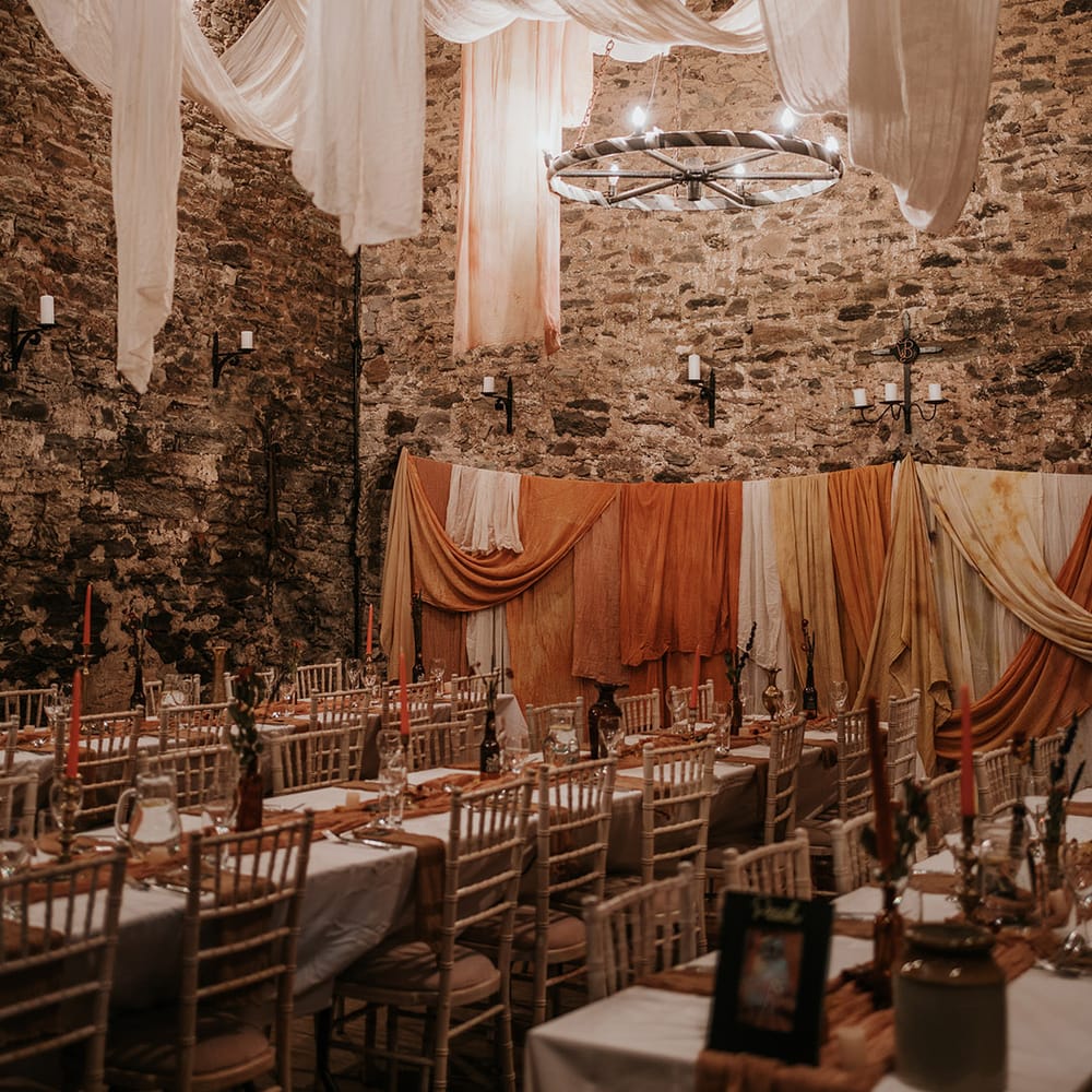 Rustic boho wedding reception room with festoon chandelier lighting, off white and burnt orange wedding drapes decorations, fairy lights and dried flower centrepieces 