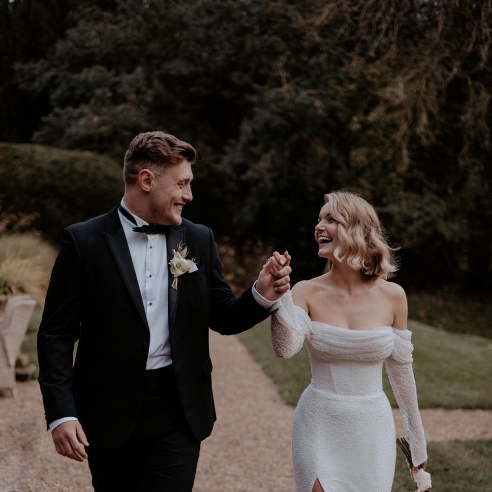 Bride in corset style wedding dress with off the shoulder long sleeves walking hand in hand with th groom in a black tuxedo 