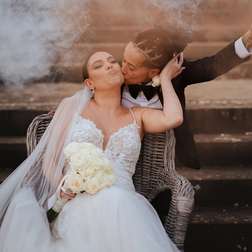 Bride in black snakeskin print tuxedo with black bowtie kissing bride on the cheek in lace sleeveless v neck Enzoani wedding dress with tulle finish during wedding smoke bomb photoshoot 