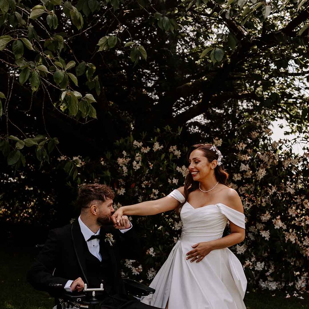 groom wearing a tuxedo in a wheelchair kissing his bride's hand in an off the shoulder wedding dress
