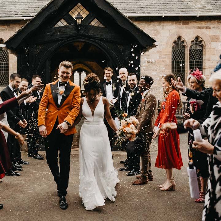 Bride in v neck sleeveless wedding dress with 3D floral applique train holding blush and orange garden roses, poppies and carnations, dried flowers and eucalyptus wedding bouquet walking with groom in crushed burnt orange velvet grooms blazer, black bowtie and black suit trousers doing colourful confetti exit