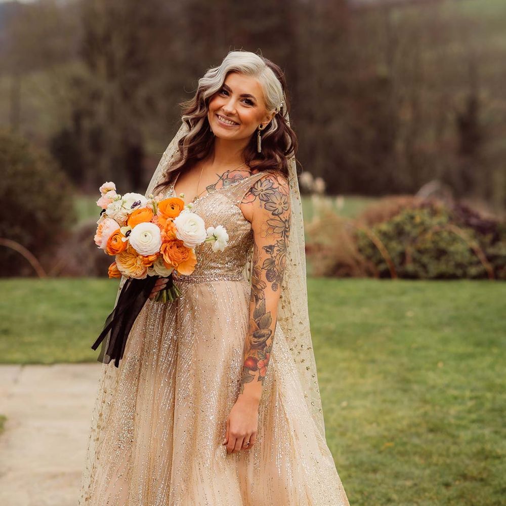 Bride in sleeveless deep v neck sparkly gold wedding dress with sheer and gold polka dot wedding veil holding orange garden rose, white poppy, light pink carnation and foliage bridal bouquet wrapped in sparkly gold ribbon at Kingscote Barn Tetbury wedding venue