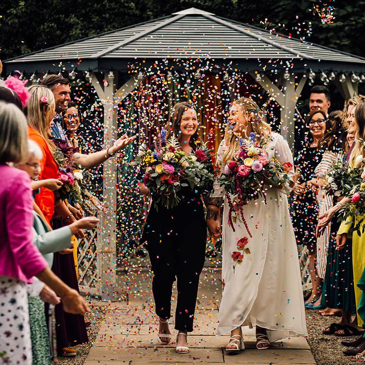 Outdoor wedding ceremony for two brides who walk back down the aisle as colourful confetti is thrown over them 