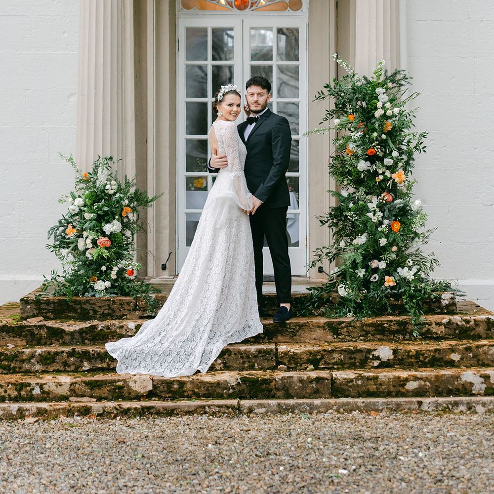 Bride in long sleeve boho lace wedding dress with groom in black tuxedo with orange flower column decorations on the stairs 