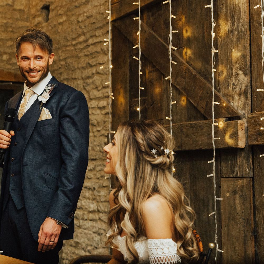 Groom smiles at bride during Groom's speech at Tythe Barn Wedding with boho decor