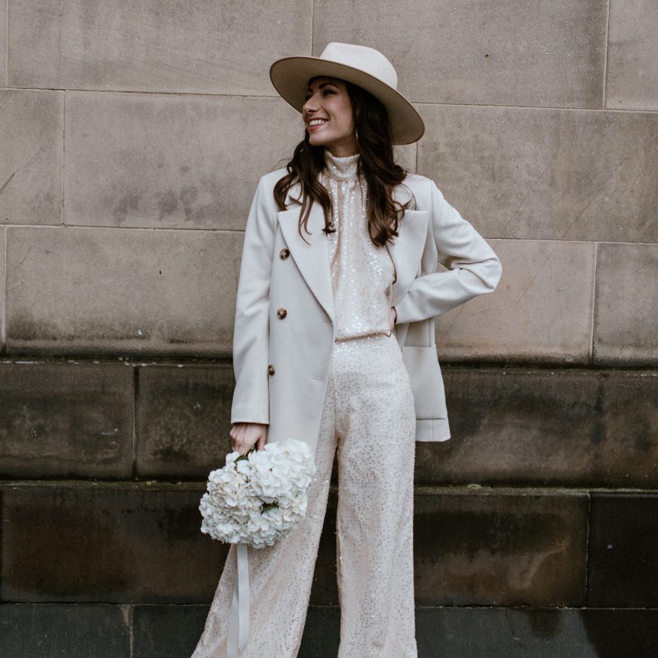 Stylish bride in wide leg sequin trousers and matching top with beige coat, white boots and Fedora hat 