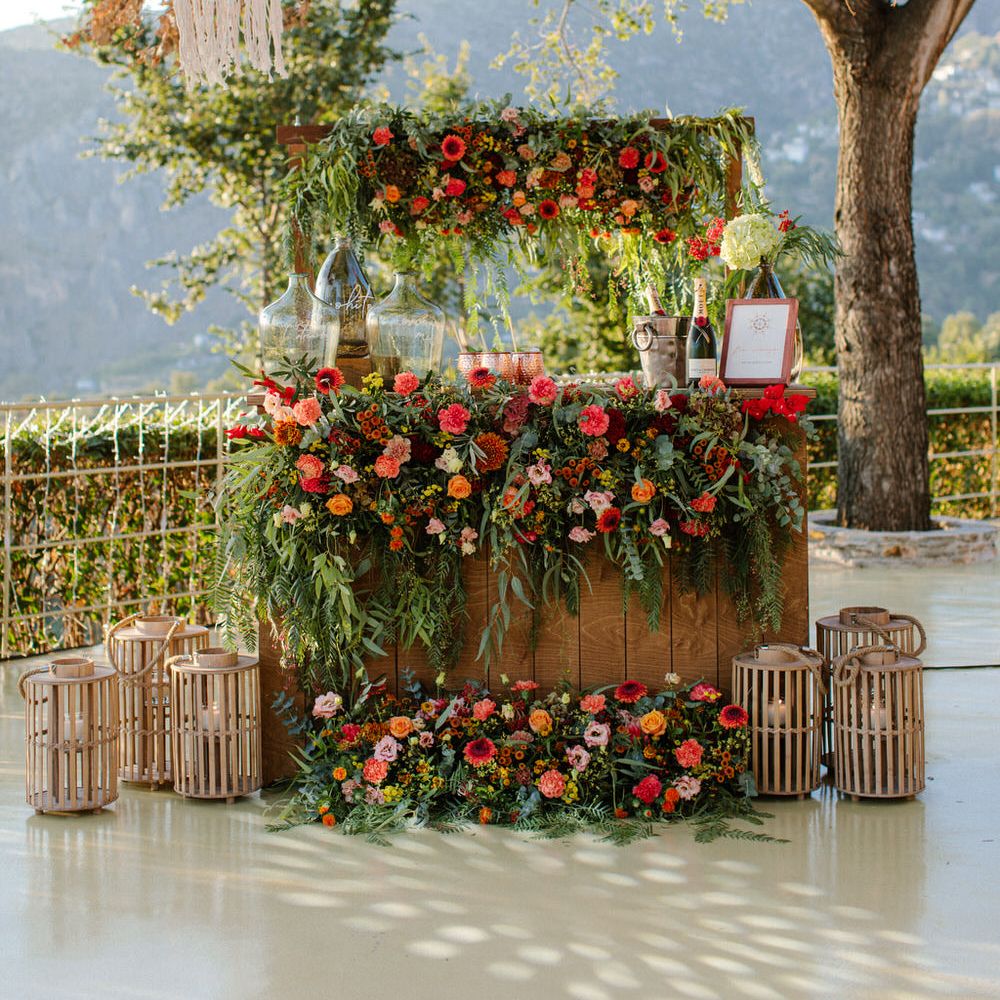 An outdoor cocktail bar covered in orange, red and pink flowers to match the bride's bouquet, with lanterns on either side