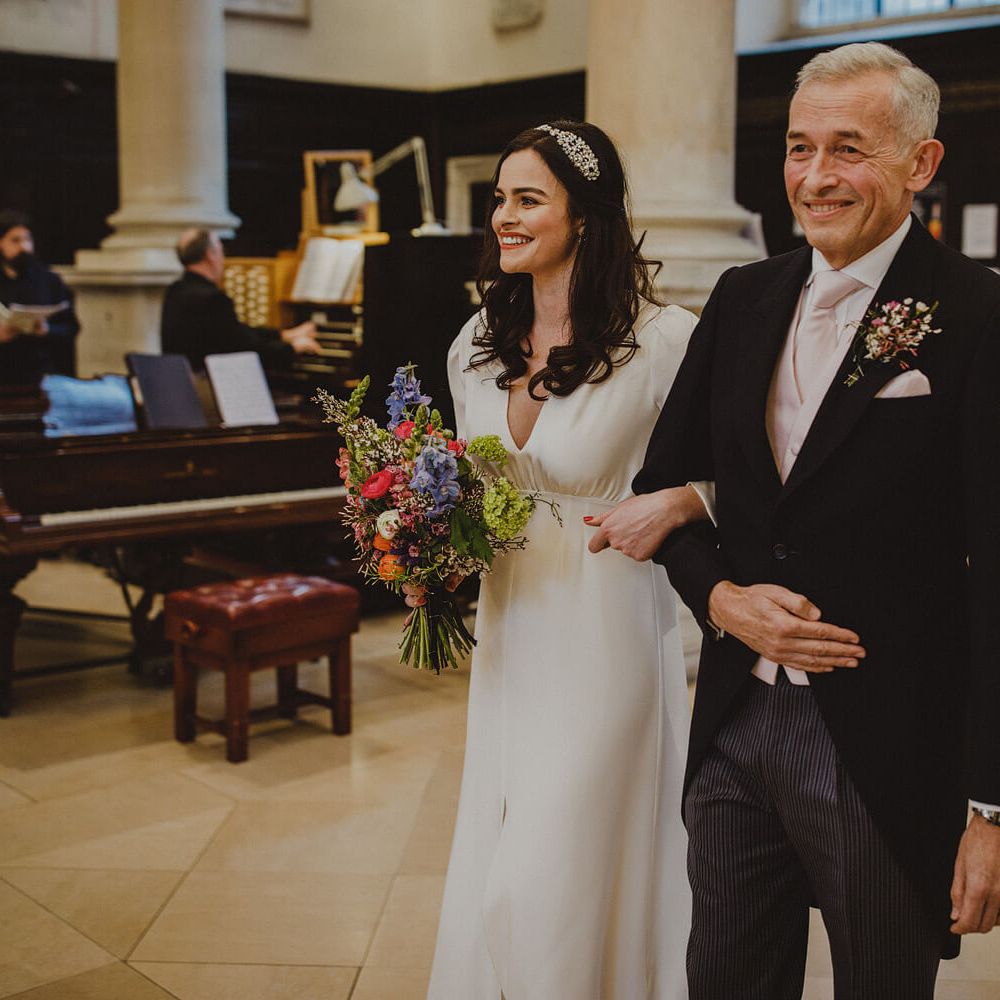 Bride walking down the aisle of London church hooking arms with older man both smiling