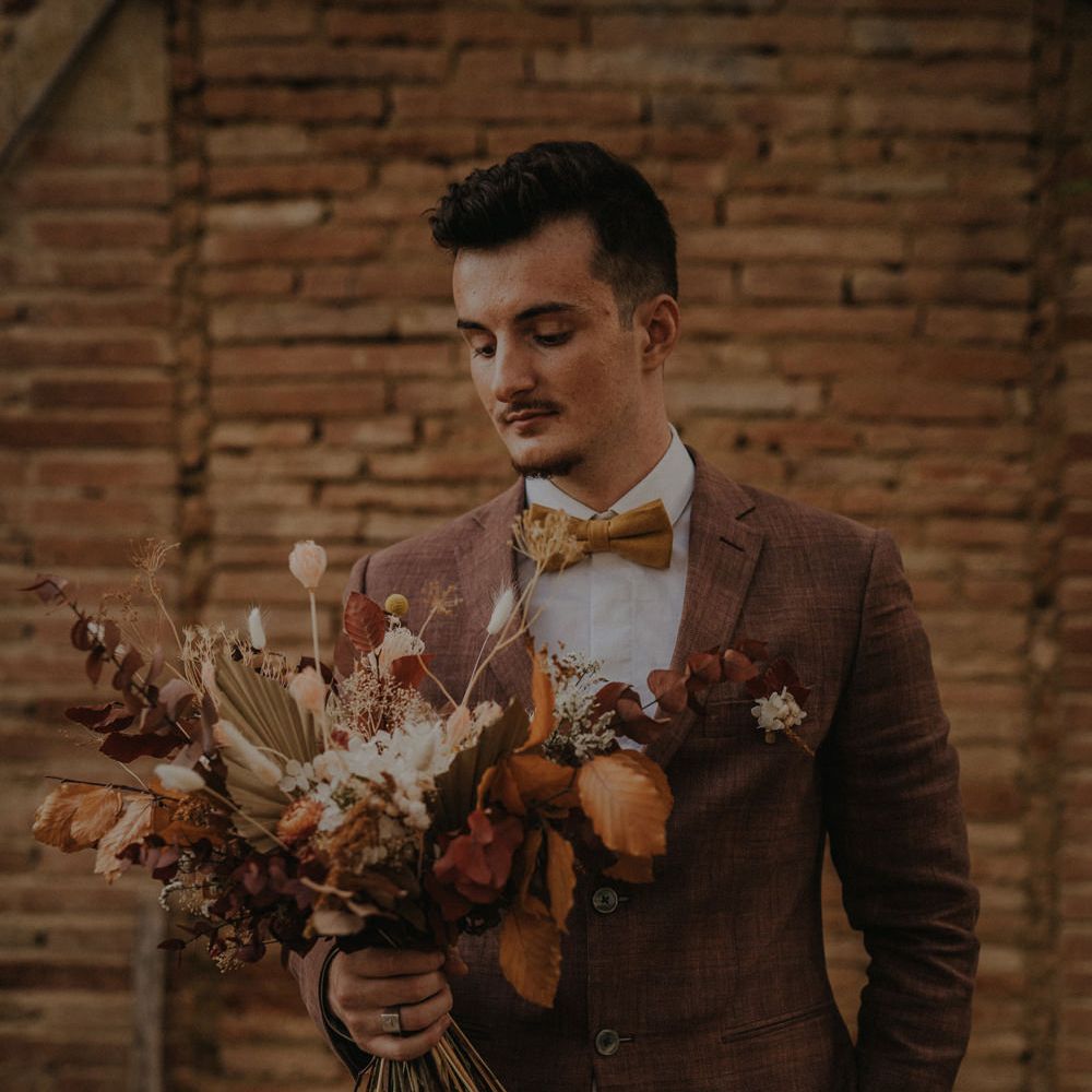 Groom in a brown wedding suit and orange bow tie holding an autumn leaf and flower wedding bouquet 