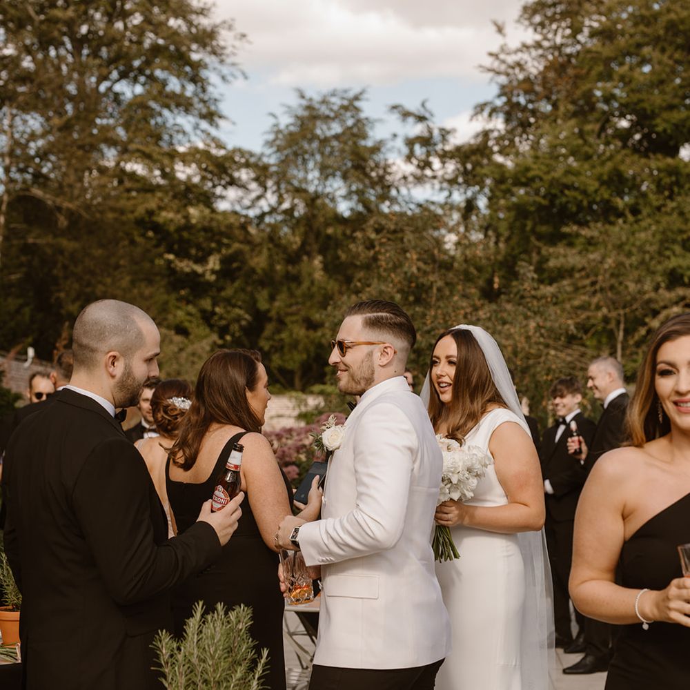 Groom in white tuxedo jacket and sunglasses and bride in Made With Love wedding dress greet guests outside the Fig House at Middleton Lodge