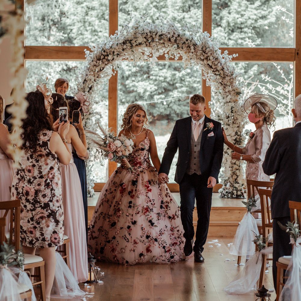 Bride & groom walk down the aisle after wedding ceremony with floral arch in the background